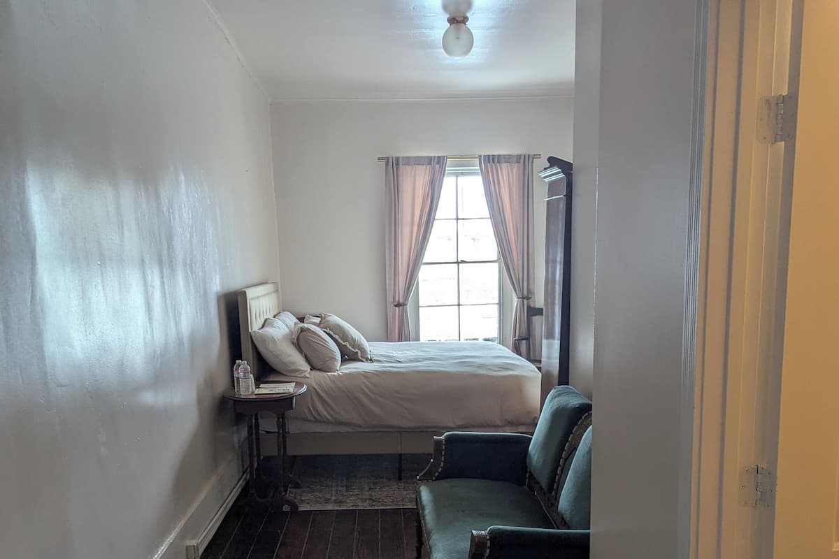 Un dormitorio con cama, mesita de noche, silla y ventana con cortinas.