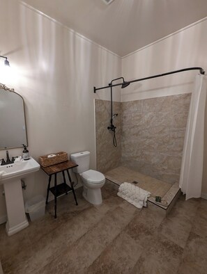 A bathroom with a shower, toilet, sink, and a small table with a basket. - Hotel Leger (Mokelumne Hill)