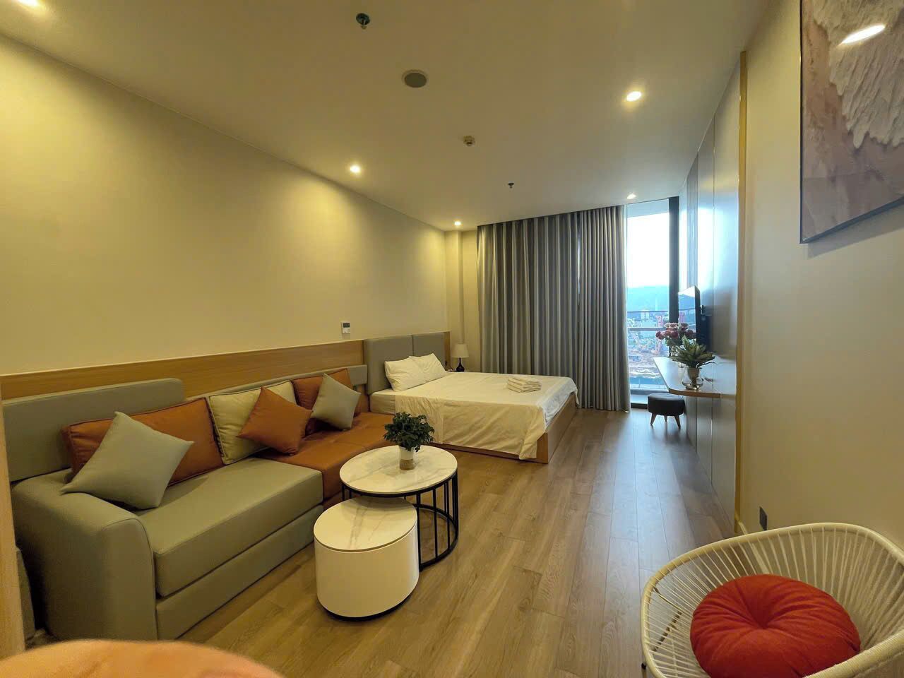 A modern hotel room with a sofa, two single beds, a round coffee table, and a view of the sea.