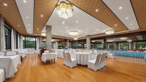 Une salle de banquet spacieuse, avec des tables rondes et des chaises blanches, de grandes fenêtres et des lustres.