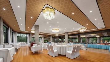 A spacious banquet hall with round tables and white chairs, large windows, and chandeliers.