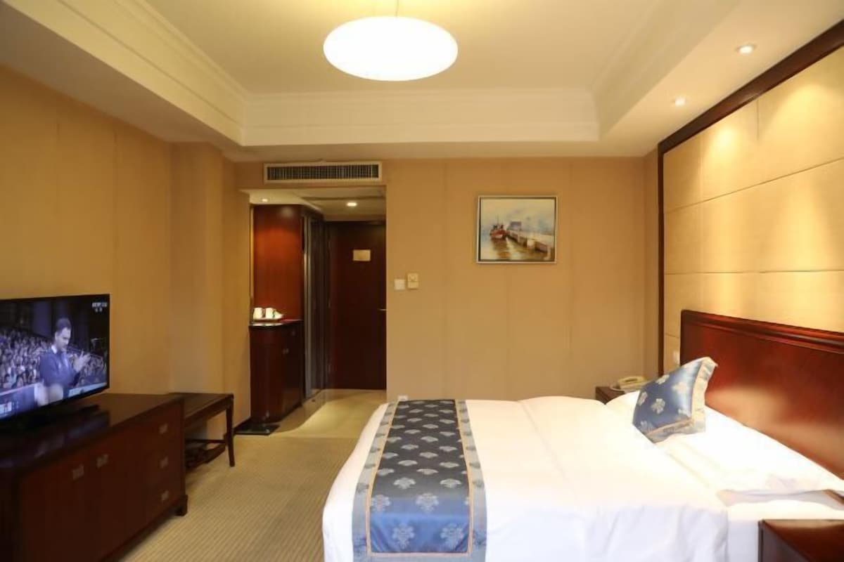 A hotel room with a television, a bed with a blue and white bedspread, a wooden headboard, a nightstand, and a painting on the wall.