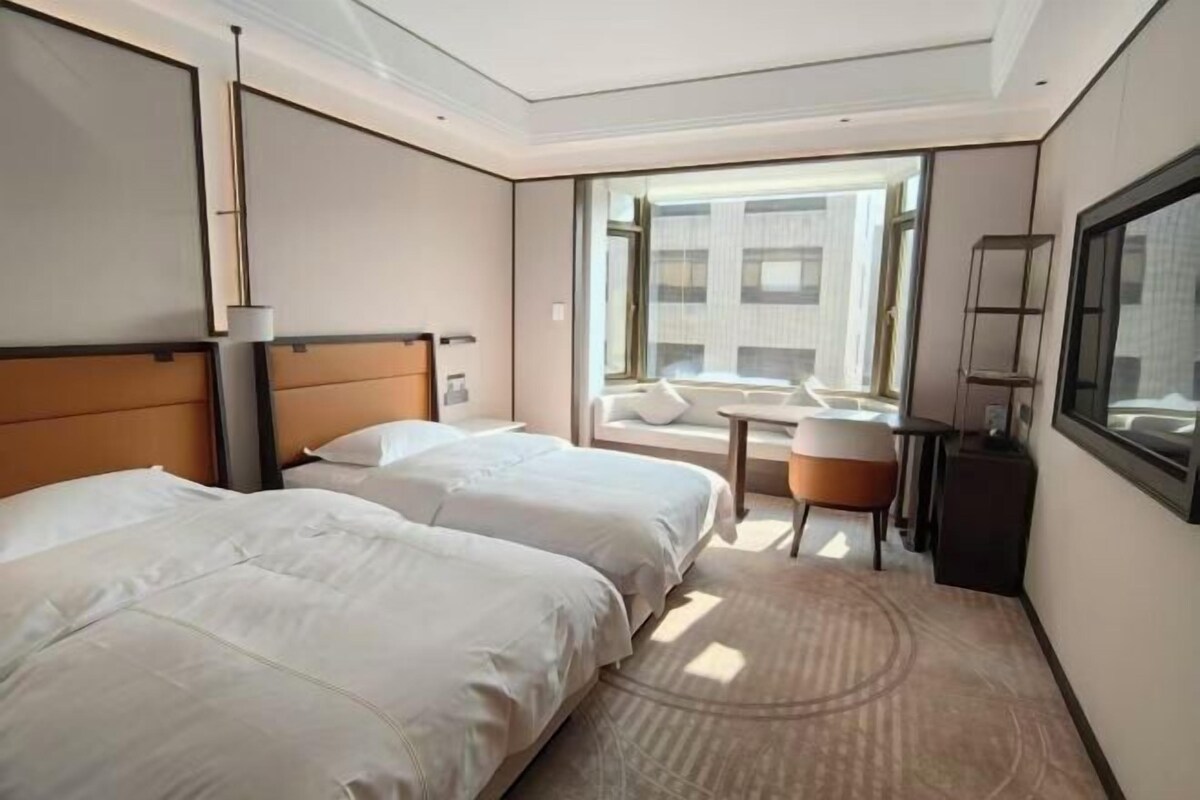 A hotel room with two beds, a small table, a chair, a desk, and a large window.