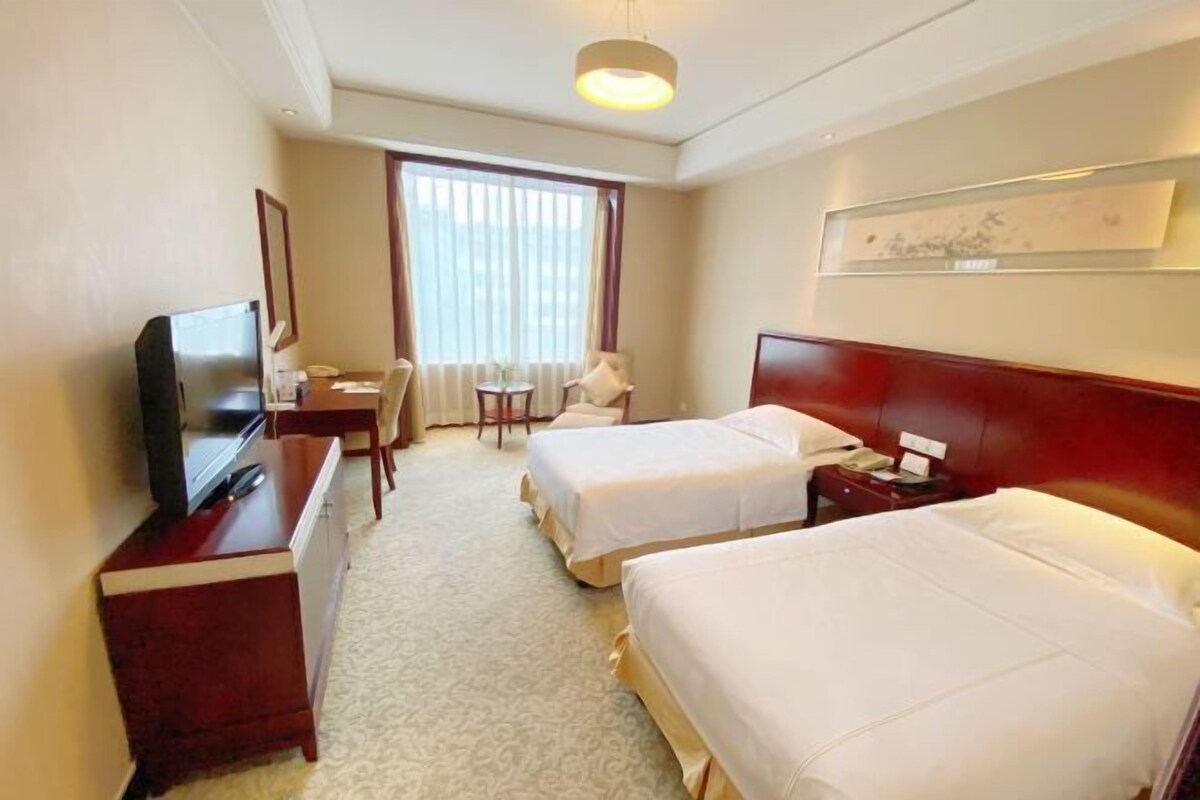 A hotel room with two beds, a desk with a TV, a chair, a small table, and a window with curtains.