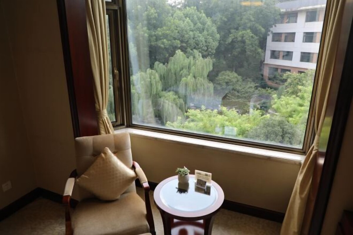 A room with a view of a lush garden through a large window, a small round table with a plant and a book, and a chair with a cushion.