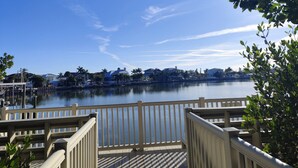 A waterfront view with a wooden deck and railing, calm water, and buildings in the background. - Boutique Beach Escape in Indian Rocks Beach - 150 steps to the sand (Indian Rocks Beach)