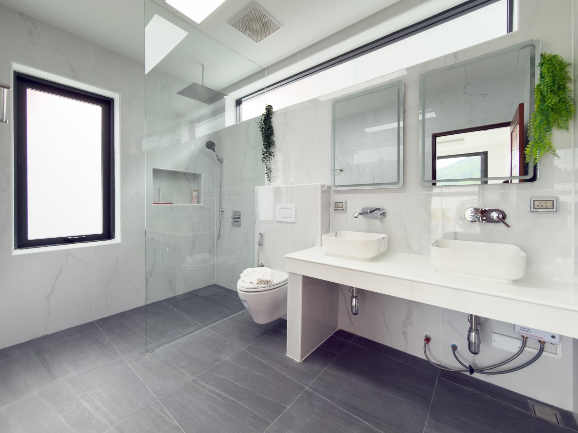 A modern bathroom with a large window, a walk-in shower, a toilet, and a vanity with two sinks.