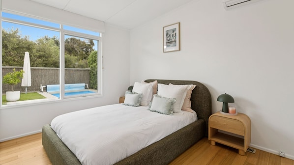 Un dormitorio con cama, mesita de noche con lámpara, cuadro en la pared y ventana con vista a una piscina.