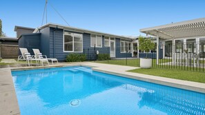 A residential swimming pool with a clear blue water surface, surrounded by a fenced yard with a covered patio area.