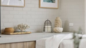 Una encimera de cocina con protector mural de ladrillo blanco, una jaula decorativa, un florero y una bandeja tejida.