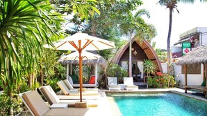 A poolside area with lounge chairs, a thatched-roof hut, and a building with a red cross sign. - Unzipp Lumbung (Gili Trawangan)
