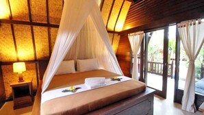 A bed with a canopy, a bedside table with a lamp, and a view through large windows. - Unzipp Lumbung (Gili Trawangan)