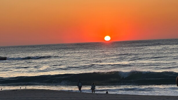 Un coucher de soleil sur l’océan, avec des vagues et quelques personnes sur la plage.