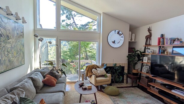 Ein geräumiges Wohnzimmer mit einer großen Couch, einem Couchtisch und einem Fernseher. Große Fenster sorgen für natürliches Licht und einen Blick ins Freie.