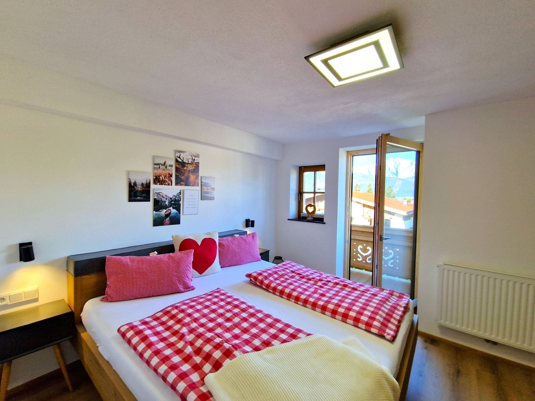 Een tweepersoonsbed met rood-witte geruite beddengoed, een nachtkastje met een lamp en een raam met uitzicht op een berg.