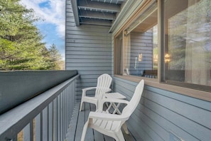 A balcony with two white plastic chairs and a table, a window with blinds, and a view of trees. - Brookside II B-205 - Sunday River Slopeside Condominium (Newry)