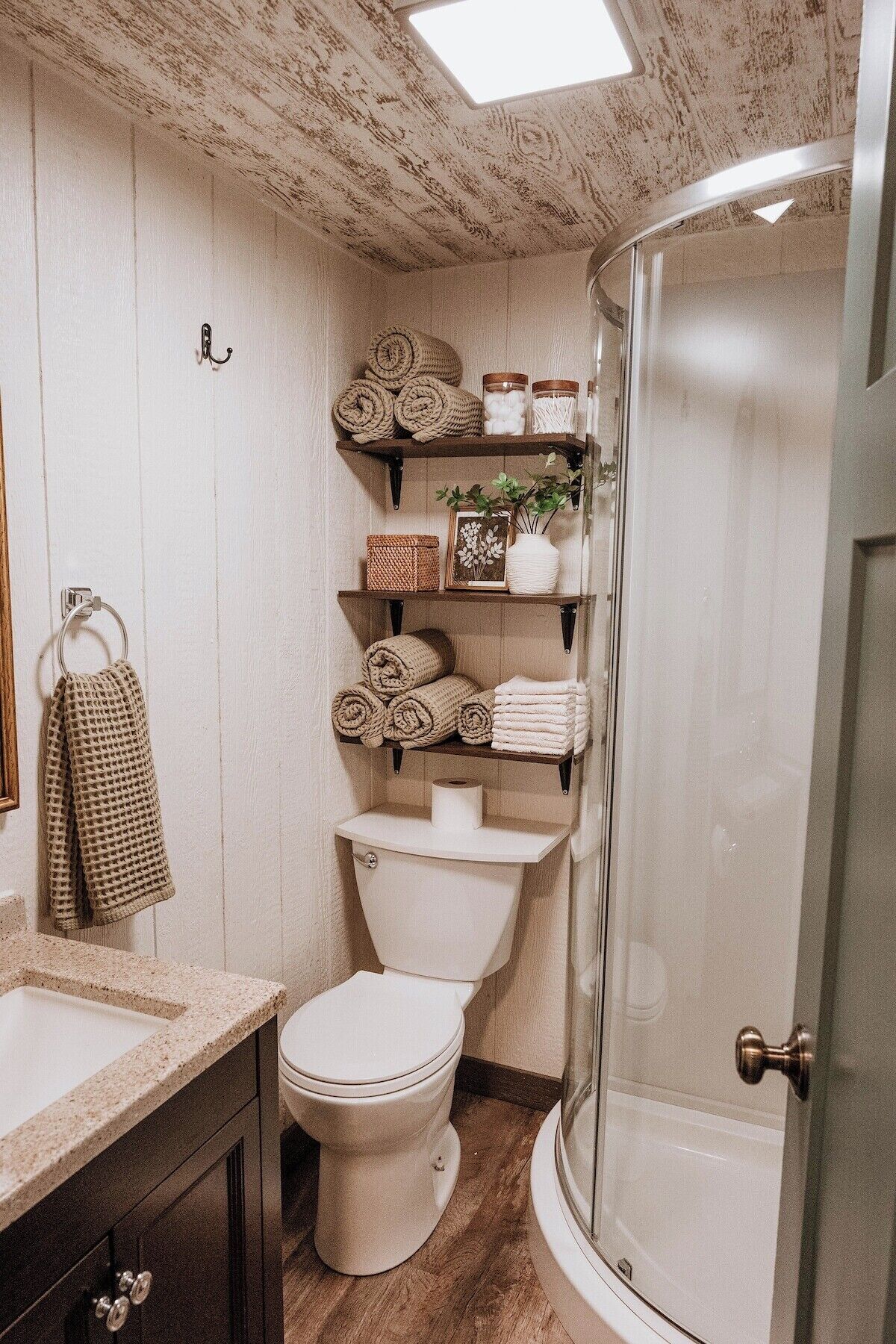A bathroom with a toilet, sink, and shower. There are shelves with towels and a framed picture.