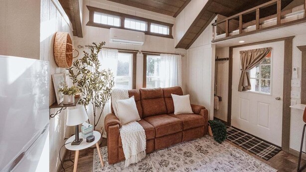 Ein gemütliches Wohnzimmer mit einem braunen Sofa, einem kleinen Beistelltisch mit Lampe, einer Topfpflanze und einem gemusterten Teppich.
