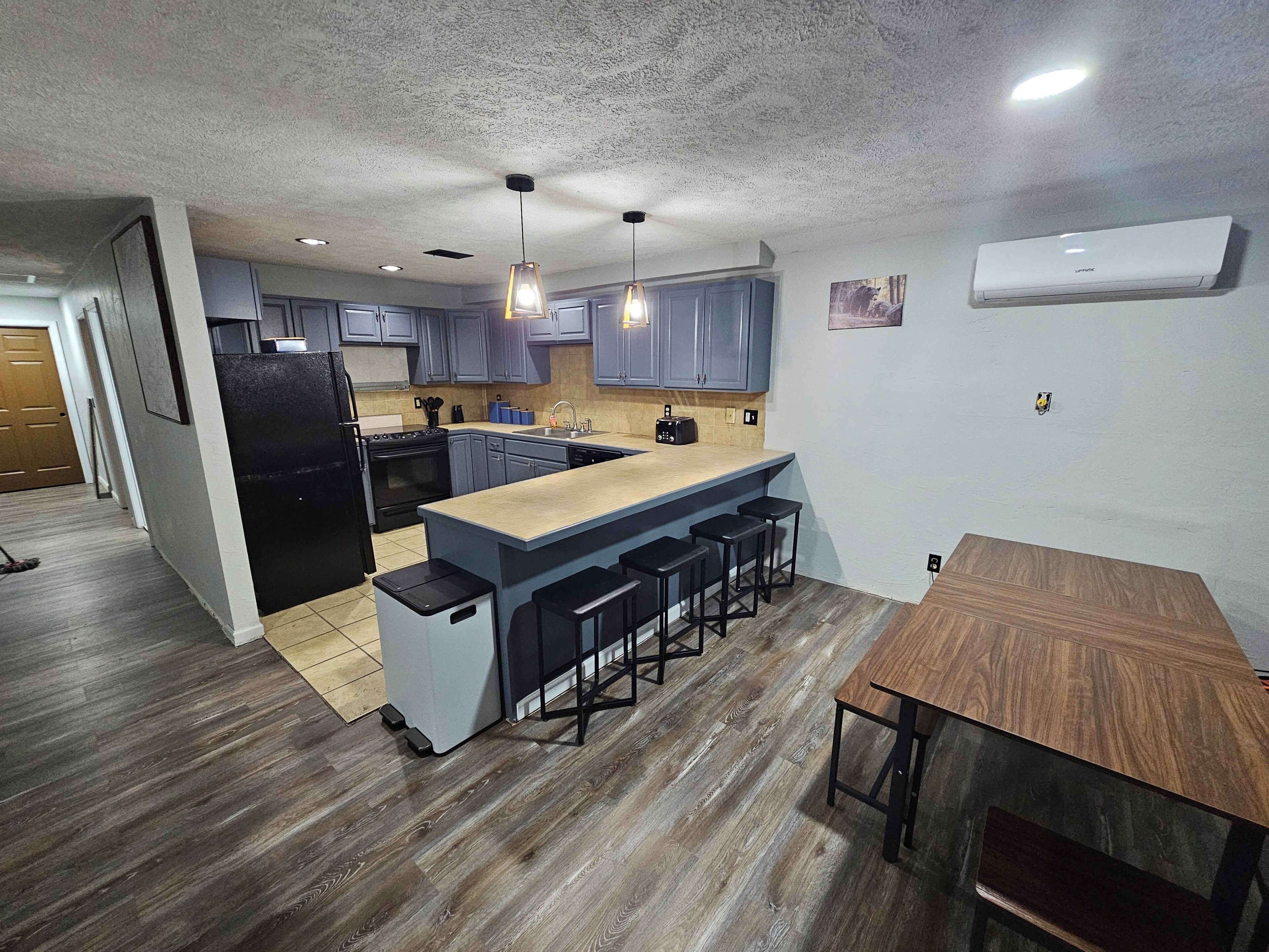 Una cocina moderna con armarios grises, un refrigerador negro, una mesa de comedor de madera y una zona con taburetes de bar.