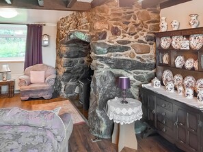 Ein gemütliches Zimmer mit Kamin aus Stein, einem bequemen Sessel, einem kleinen Tisch mit Lampe und einer Vitrine mit Dekorationsplatten.