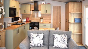 Un espacio habitable compacto con kitchenette, sofá gris con cojines estampados, muebles de madera y una pequeña mesa de comedor.