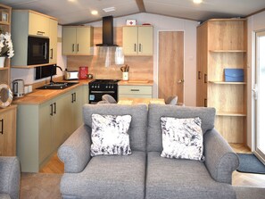 A compact living space with a kitchenette, grey sofa with patterned cushions, wooden cabinets, and a small dining table. - This three-bedroom lodge at Beverley Holiday Park, Paignton. (Paignton)