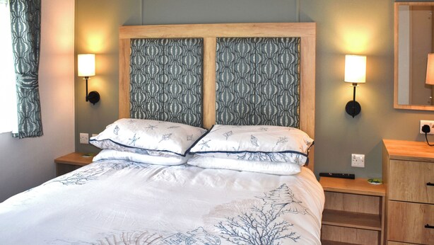 Una cama bien tendida con una colcha estampada, dos lámparas fijadas a la pared y una mesita de noche de madera con un cajón.