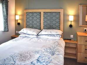 A neatly made bed with a floral bedspread, two wall-mounted lamps, and a wooden nightstand with a drawer. - This three-bedroom lodge at Beverley Holiday Park, Paignton. (Paignton)