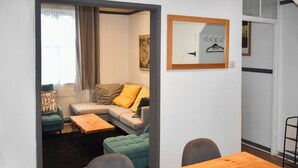 Ein modernes Wohnzimmer mit einer Couch, zwei Sesseln, einem Couchtisch und einem Esstisch. An der Wand hängt ein Gemälde, und ein Kleiderständer steht in der Ecke.