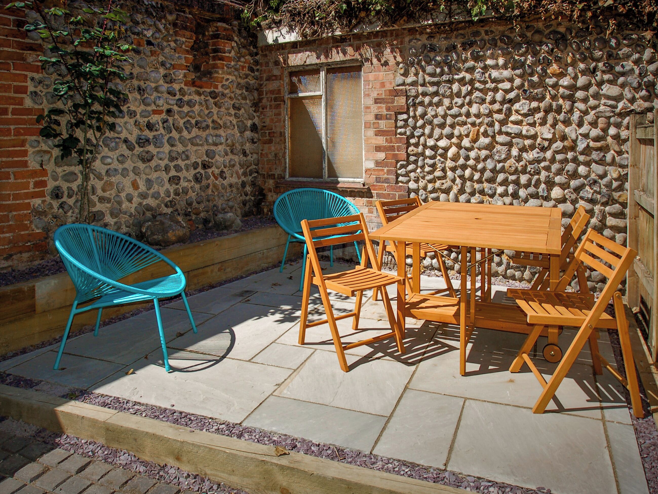 An outdoor patio with a wooden table and chairs, a stone wall, and a window with a metal screen.