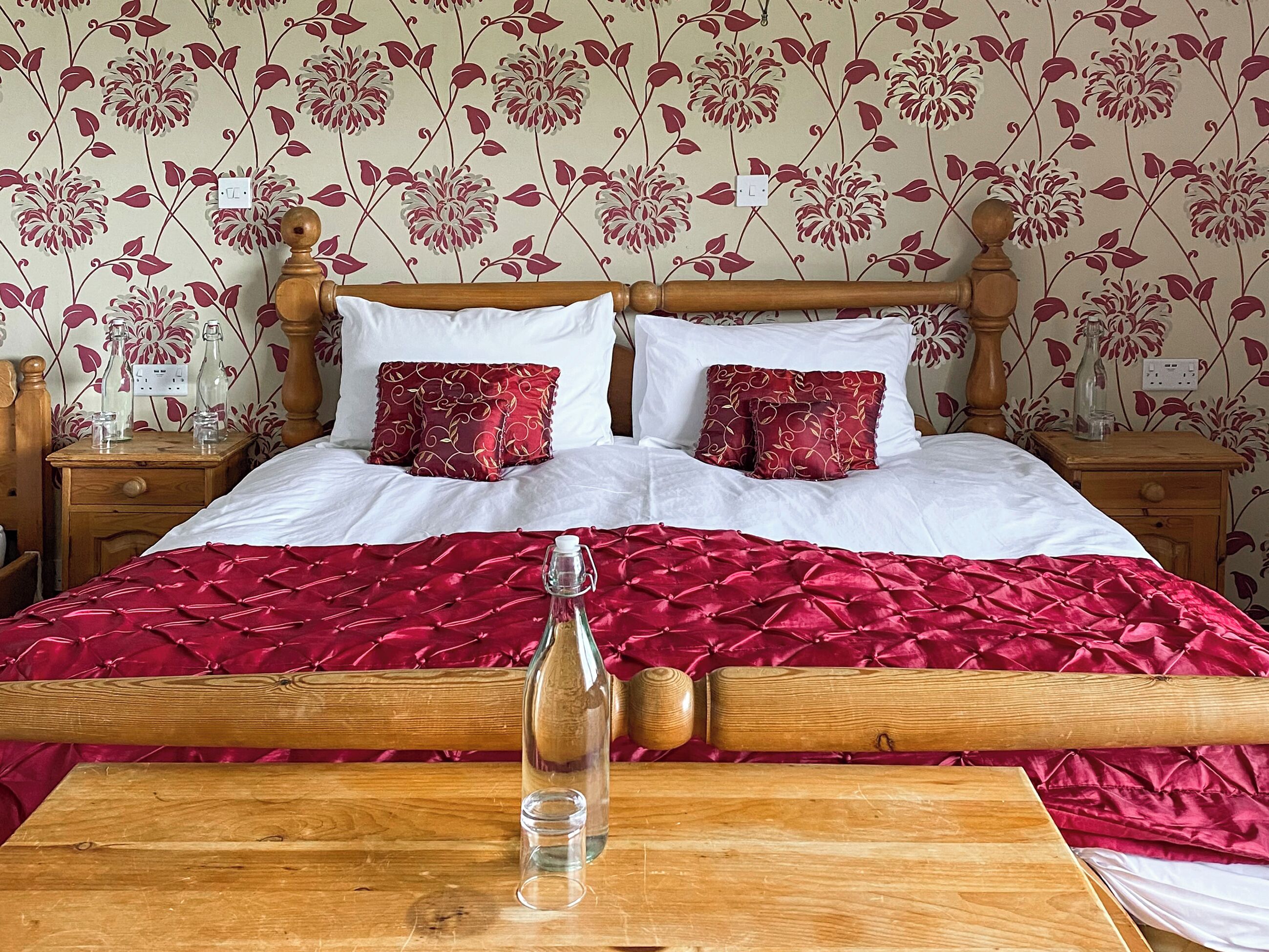 A bed with a red velvet cover, two wooden nightstands, a bottle, and a glass on a wooden table.