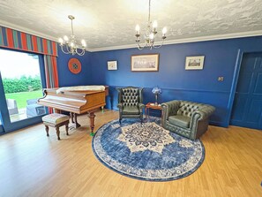A room with a grand piano, a green leather armchair, a round blue and white rug, and a chandelier. - 8 bedroom accommodation in Dingwall (Dingwall)