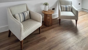 Et værelse med to stribede lænestole, et lille træ sidebord med en vase og en plante, samt et skråt loft med naturligt lys.