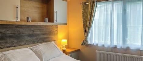 Una cama bien hecha con una bandeja de desayuno, una lámpara de noche y una ventana con cortinas translúcidas.