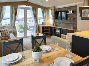 A modern living room with a fireplace, a dining area with a table set for two, and a large window offering a view of the outside. - 2 bedroom accommodation in Filey (Filey)