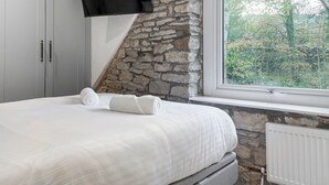 Un dormitorio con pared de piedra, una cama con ropa de cama blanca, un televisor de pantalla plana montado sobre la cama y una ventana con vista a árboles.