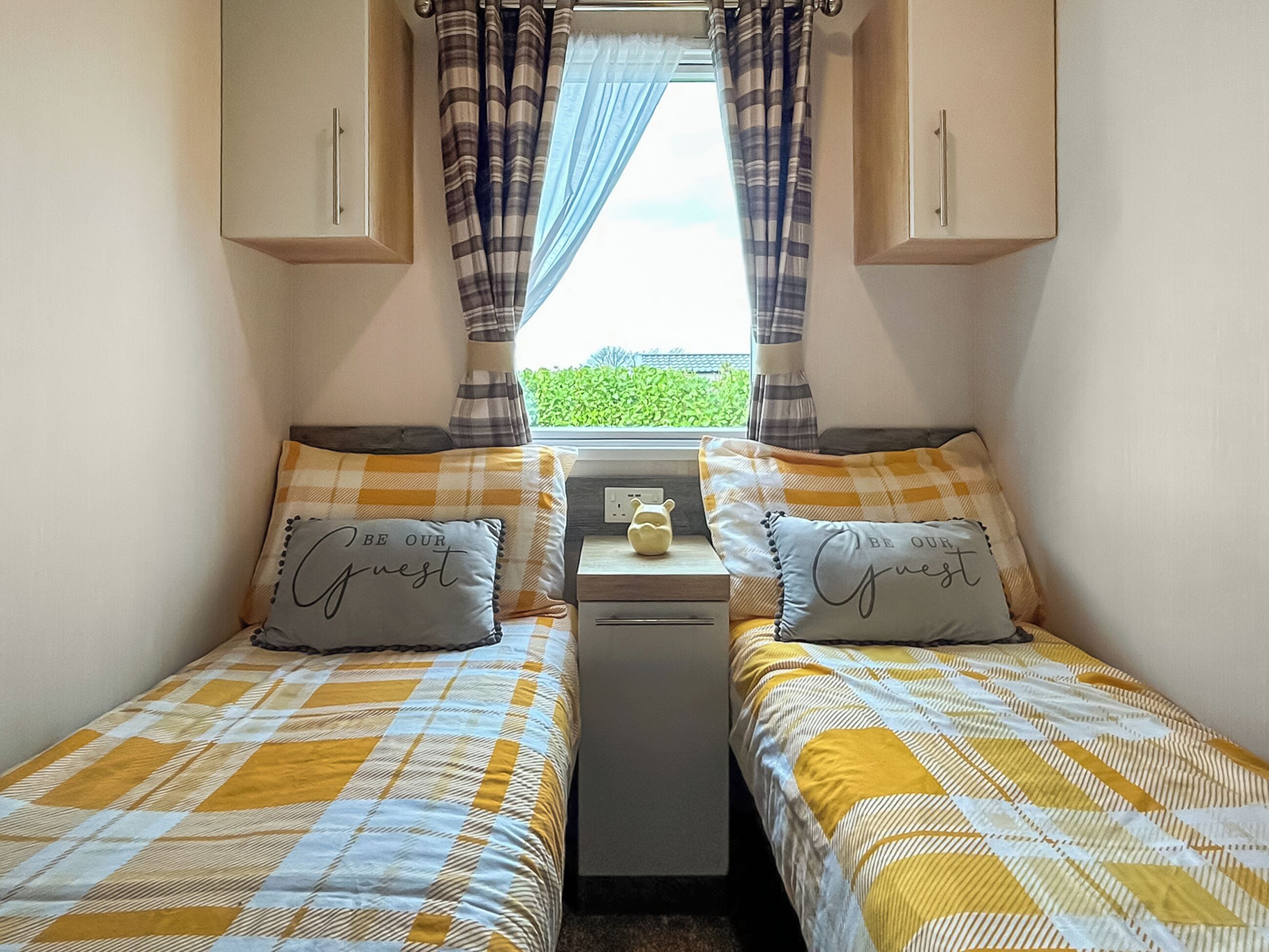 Habitació petita i acollidora amb dos llits, cada un amb un llençol a quadres i coixins amb la frase “BE OUR Guest”. Hi ha una taula petita amb una tetera i una tassa, i cortines a la finestra.