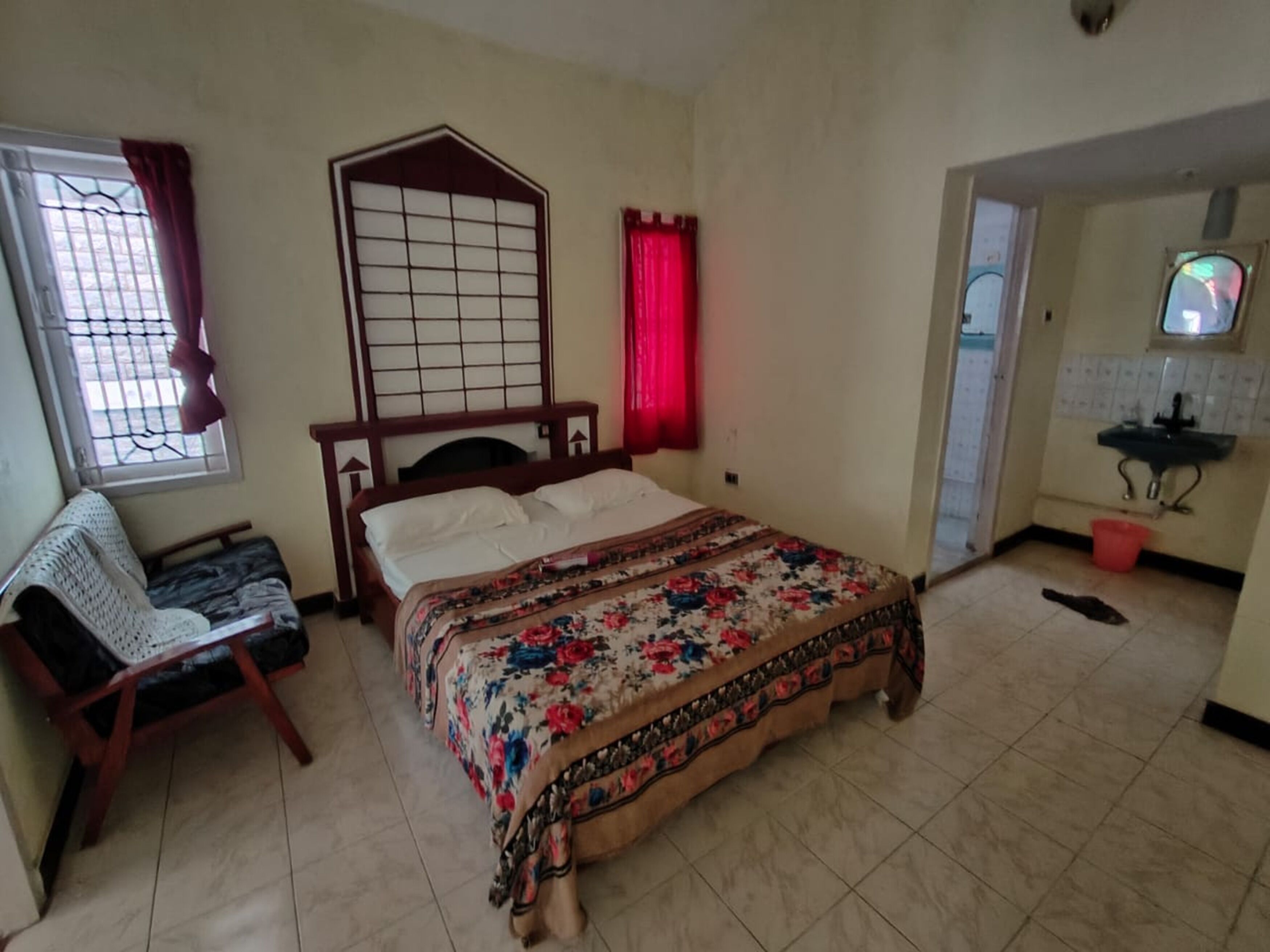 Un dormitorio con cama, una silla, ventana con cortinas, baño con lavamanos y espejo.