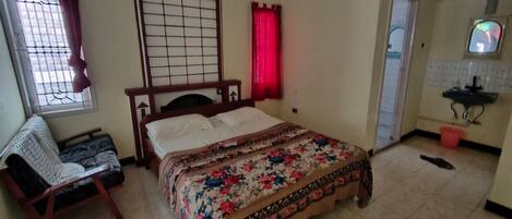 Un dormitorio con cama, una silla, ventana con cortinas, baño con lavamanos y espejo.