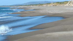 Uma praia com areia e água, céu claro e colinas ao longe.