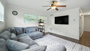 A living room with a sectional sofa, a ceiling fan, a TV mounted on the wall, and a window with blinds. - Spacious 3BR Home w/ Kitchen, Backyard & Parking (Baldwin)