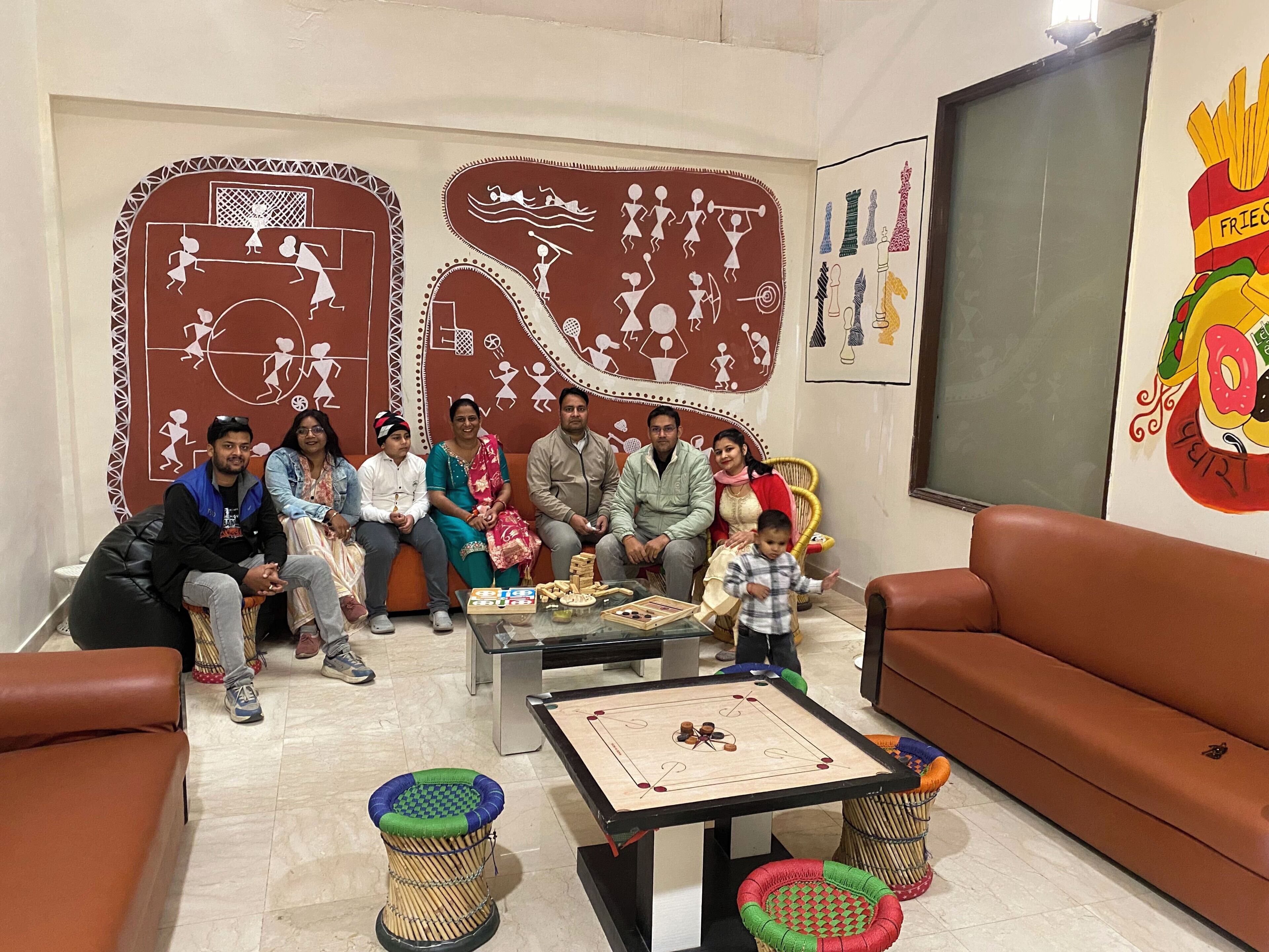 A group of people seated on a couch and chairs in a room with a checkered floor, a table with a board game, and a wall with various drawings.
