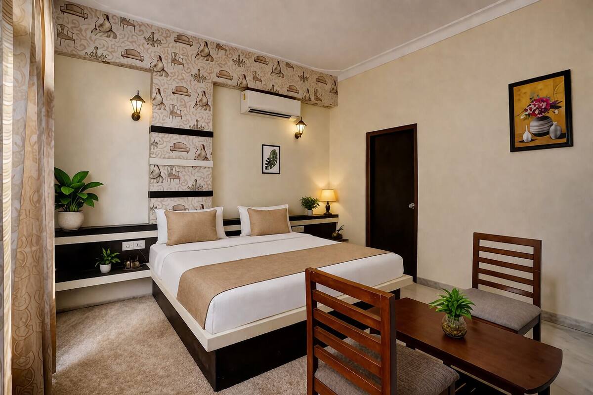 A hotel room with a bed, bedside tables, a desk, chairs, a potted plant, and a painting on the wall.