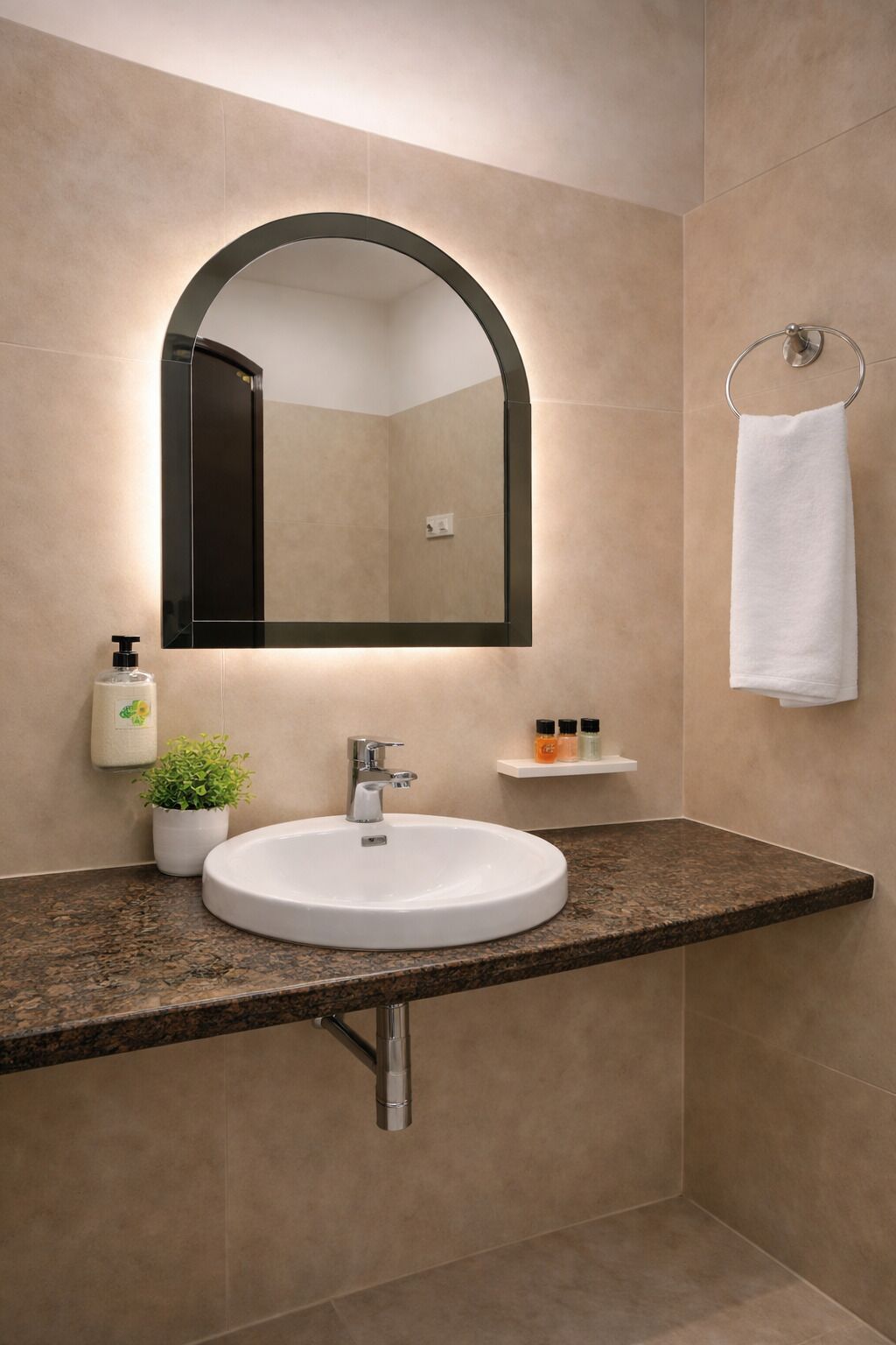 Une salle de bain avec un lavabo rond, un miroir avec un cadre en arc de cercle, un porte-serviettes, un distributeur de savon et une petite étagère contenant des articles de toilette.