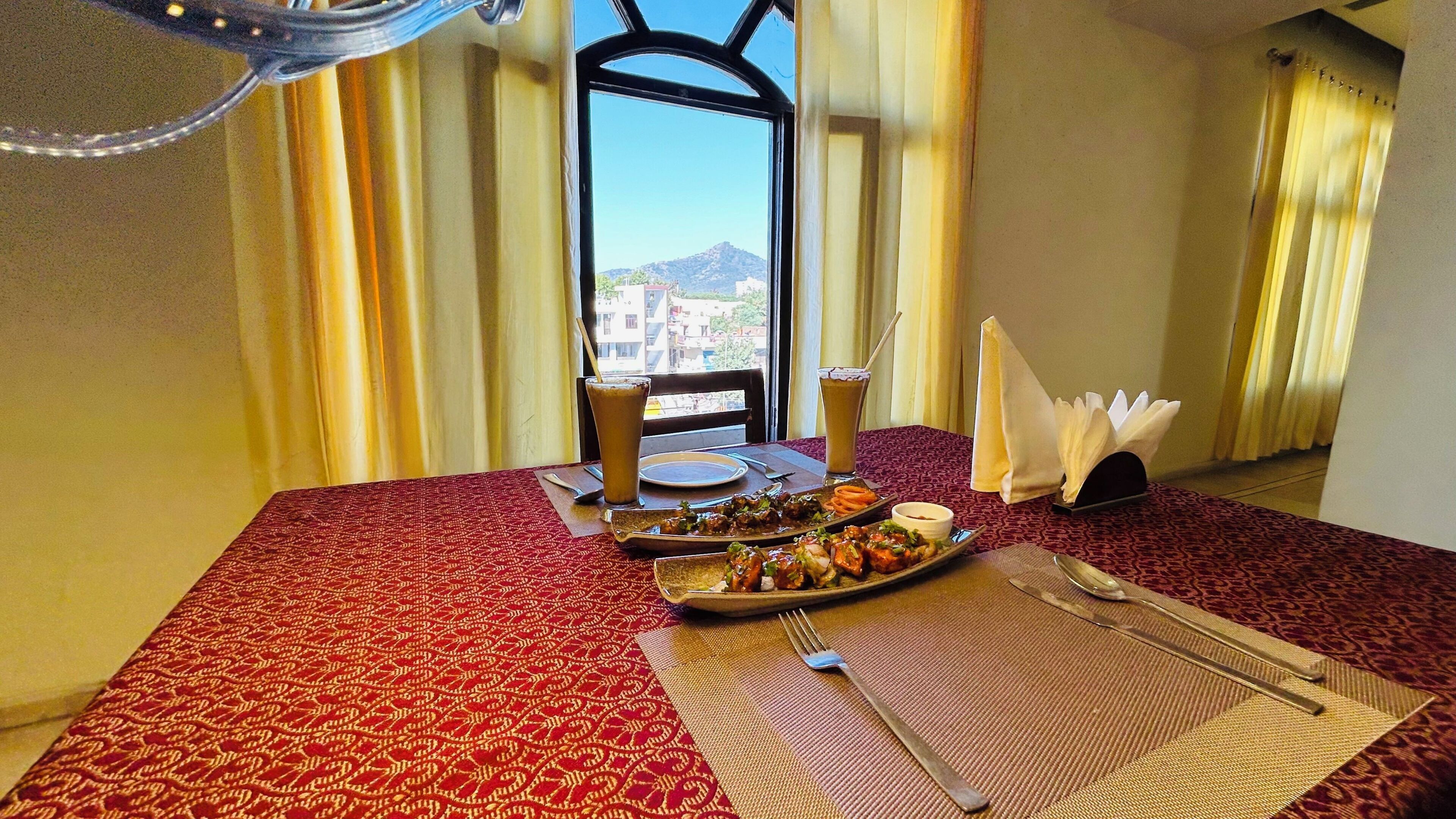 A hotel room with a dining table set for two, a view of buildings and mountains through the window, and a red patterned tablecloth.