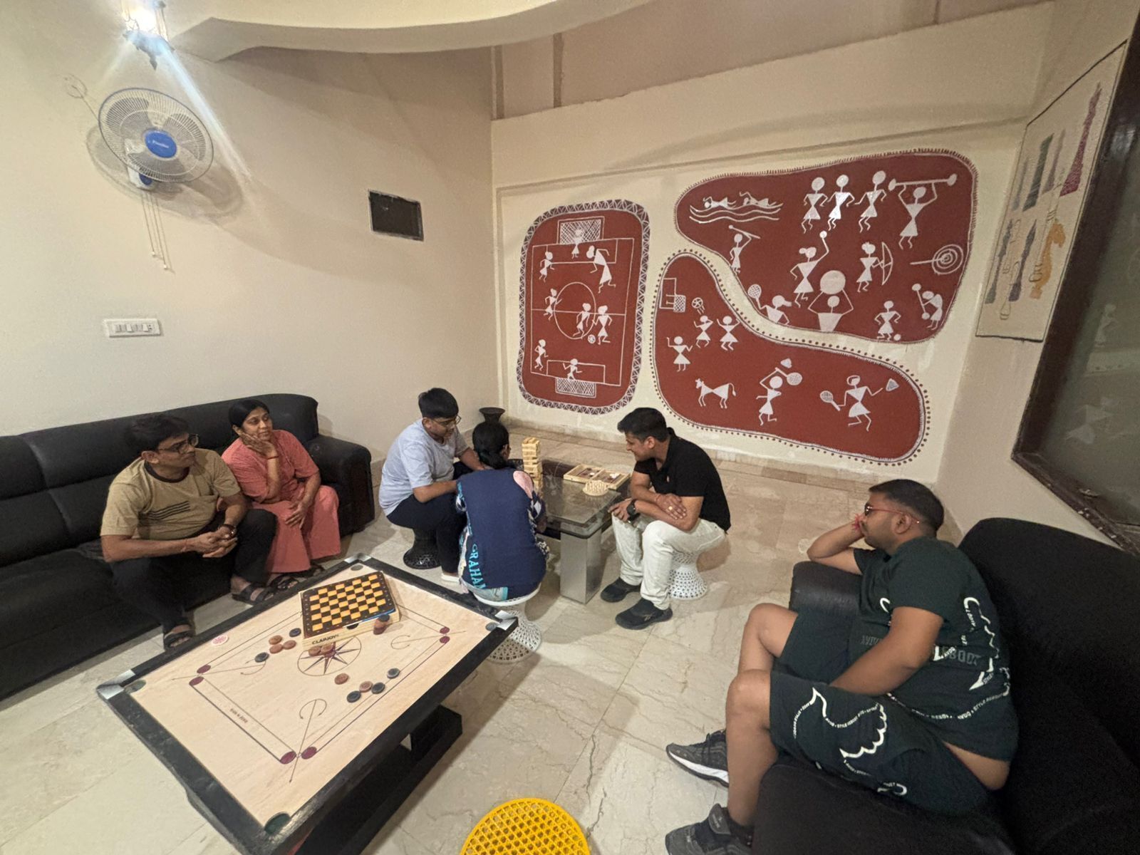 Un grupo de personas jugando carrom en una habitación con un gran mural y un ventilador.
