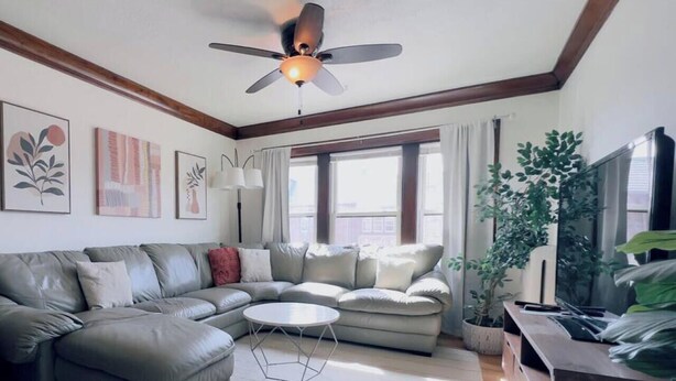 A modern living room with a sectional sofa, a ceiling fan, large windows, and a TV. - Babich Building - 3 Bedroom - Upper Apt #4 (West Allis)