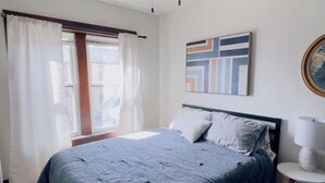 A bedroom with a bed, a ceiling fan, a window with curtains, and a wall-mounted abstract painting. - Babich Building - 3 Bedroom - Upper Apt #4 (West Allis)
