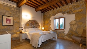 Une chambre avec un mur en pierre, des poutres en bois au plafond, un lit avec du linge de maison blanc, deux lampes de chevet et une fenêtre donnant sur un paysage verdoyant.