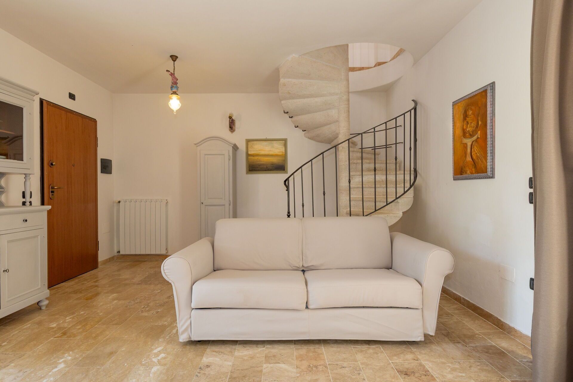 Een kamer met een witte bank, een spiltrap, een radiator en een deur.